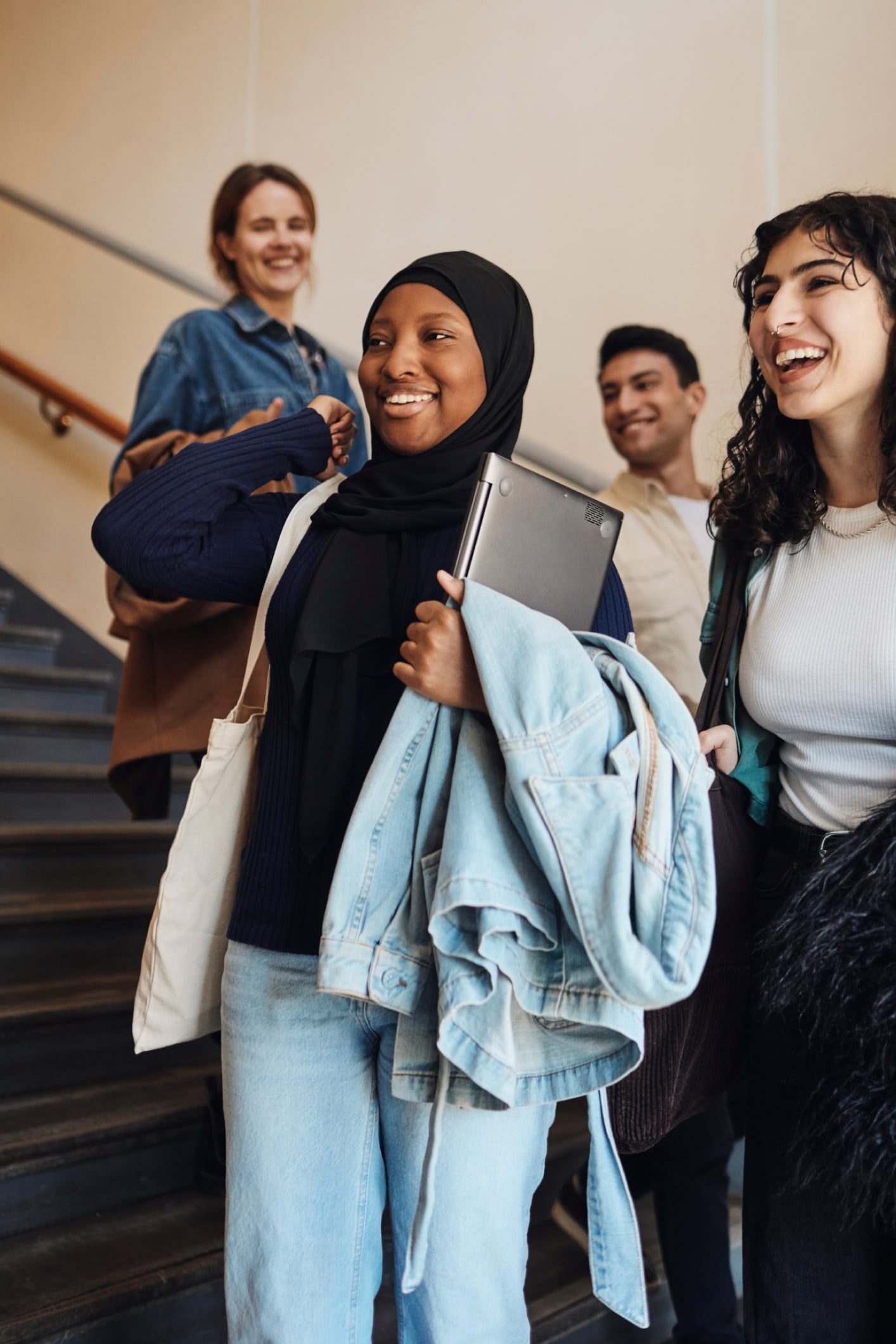 Smiling woman in hijab is holding a laptop while walking down stairs