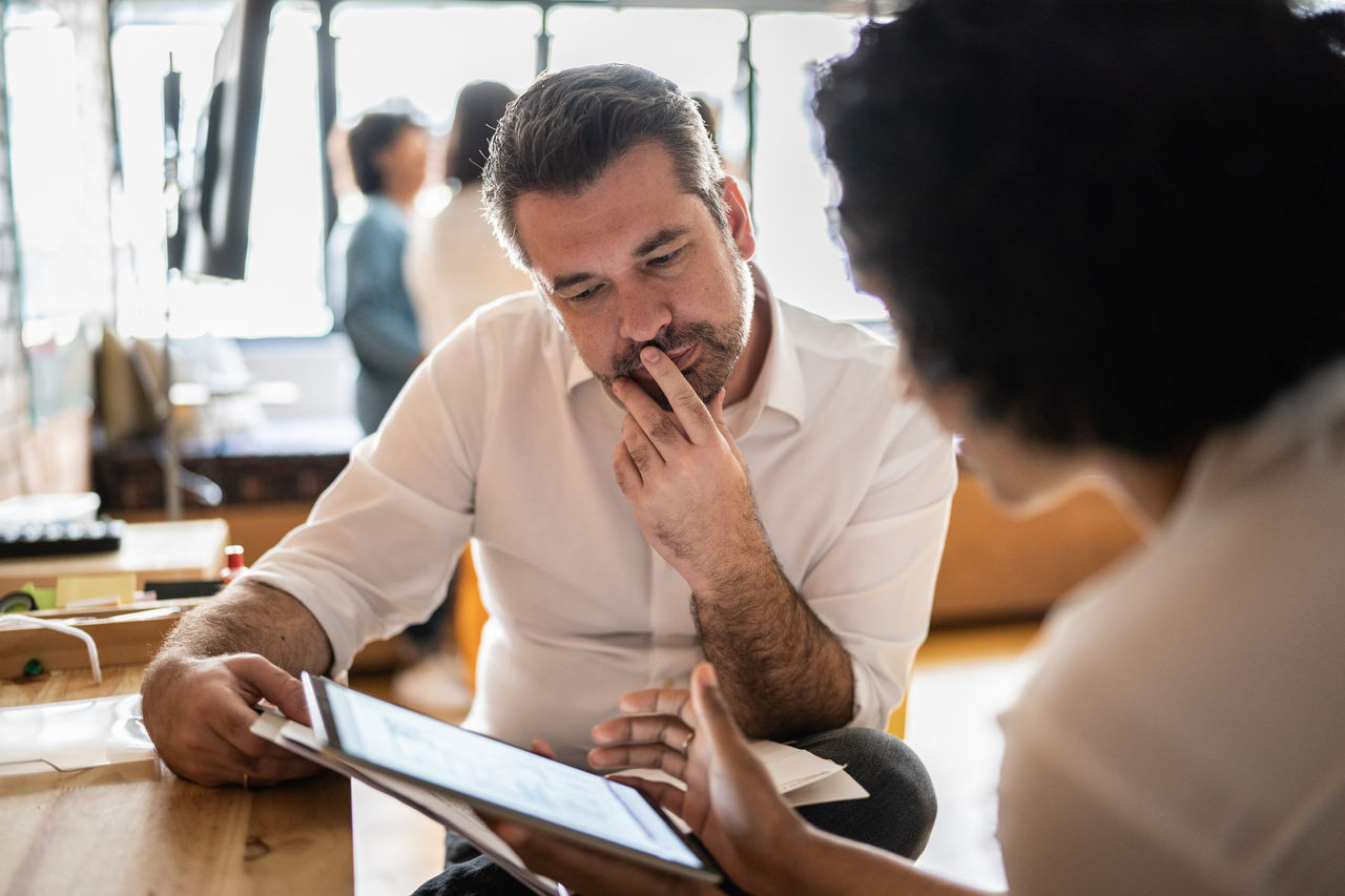 A Black woman is using a tablet screen to assist her colleague, who listens thoughtfully with his hand to his chin. 